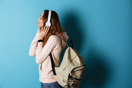 Side view portrait of a smiling happy girl student with backpack listening to music with headphones isolated over blue backgroundの写真素材