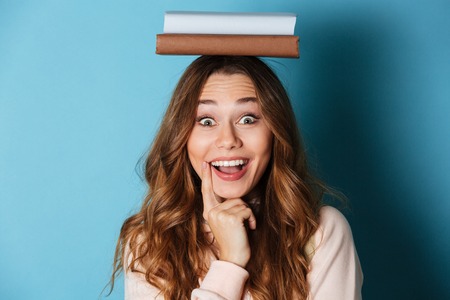 Portrait of a happy cheery girl holding books on her head and looking at camera isolated over blue backgroundの写真素材
