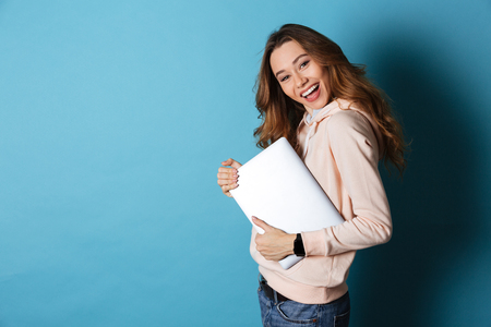 Portrait of a smiling pretty girl holding laptop computer while standing and looking at camera isolated over blue backgroundの写真素材