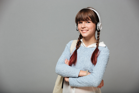 Portrait of a smiling young schoolgirl with backpack listening to music with headphones and looking at camera isolated over gray backgroundの写真素材
