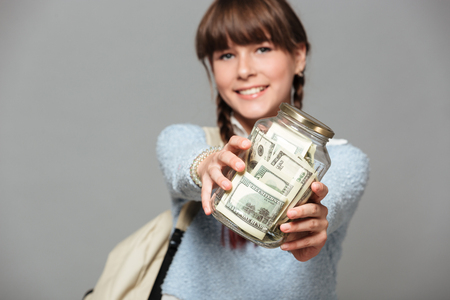 Picture of smiling girl standing isolated grey background with jar full of money. Looking camera holding backpack.の写真素材