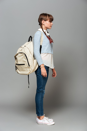 Full length side view portrait of a pretty schoolgirl with backpack standing isolated over gray backgroundの写真素材