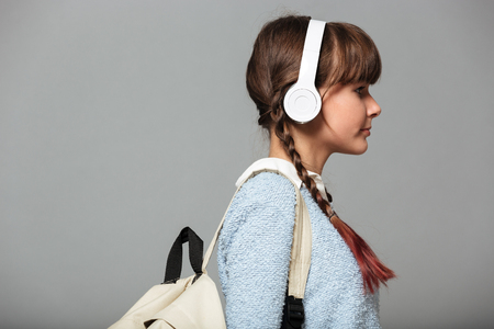 Side view photo of girl standing isolated grey background listening music with headphones. Looking aside holding backpack.の写真素材