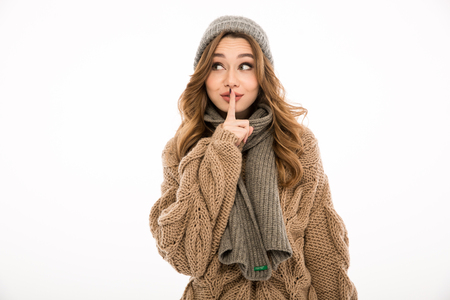Smiling young cute woman dressed in warm sweater and scarf standing isolated over white wall background. Looking aside showing silence gesture.の写真素材
