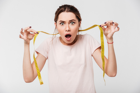 Portrait of a shocked young girl with open mouth holding measuring tape and looking at camera isolated over white backgroundの写真素材