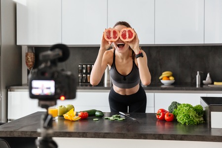 Joyful healthy young girl recording her video blog episode about healthy food diet while standing at the kitchen at home and sowing ok gestureの写真素材