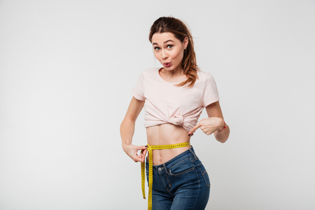 Portrait of a slim pretty woman holding measuring tape around her waist and pointing finger isolated over white background