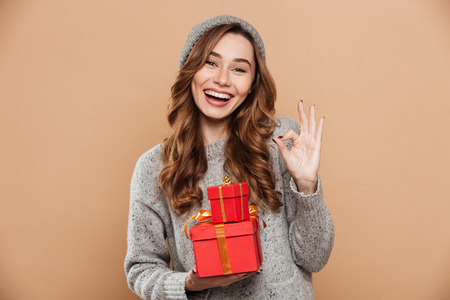 Portrait of gorgeous happy woman in warm clothes showing OK gesture while holding presents, looking at camera, isolated on beige backgroundの写真素材