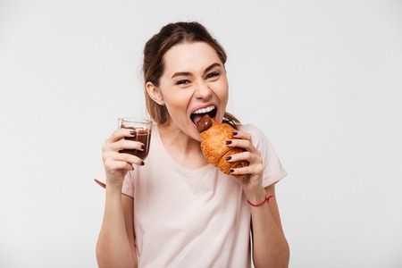 Portrait of a hungry pretty girl eating croissant and chocolate pudding from a glass isolated over white backgroundの写真素材