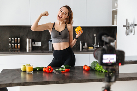 Cheerful healthy young girl recording her blog episode about healthy food diet while standing at the kitchen at home and flexing bicepsの写真素材