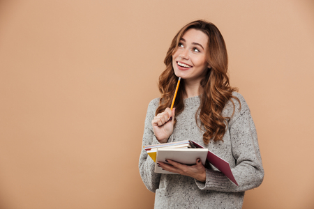 Portrait of happy thinking female student in warm sweater holding pencil and notebooks, looking aside, isolated over beige backgroundの写真素材
