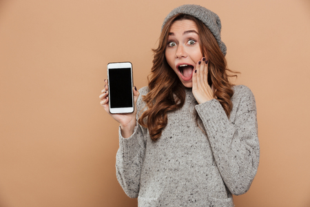 Close-up photo of amazed charming brunette woman in warm hat and knitted sweater showing blank mobile screen and looking at camera, isolated on beige backgroundの写真素材