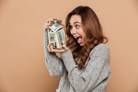 Overjoyed curly brunette girl holding glass jar with her savings, isolated over beige backgroundの写真素材