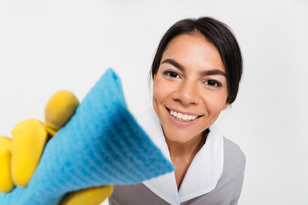 Close-up photo of young smiling housekeeper in yellow rubber gloves cleaning window with rag, isolated over white backgroundの写真素材