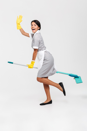 Full length photo of happy funny girl in uniform riding mop as a witch, isolated over white backgroundの写真素材