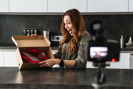 Happy young girl recording her video blog episode about new high stiletto shoes while sitting at the kitchen table at homeの写真素材