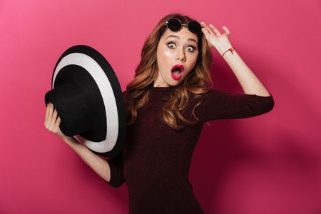 Photo of young beautiful shocked lady holding hat and wearing sunglasses standing isolated over pink background. Looking camera.の写真素材