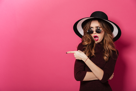 Image of young emotional lady wearing hat and sunglasses standing isolated over pink background. Looking aside pointing.の写真素材
