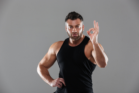 Portrait of a confident strong male bodybuilder standing and showing ok gesture isolated over gray backgroundの写真素材