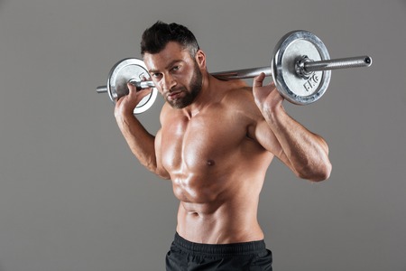 Portrait of a concentrated strong shirtless male bodybuilder holding heavy barbell isolated over gray backgroundの写真素材