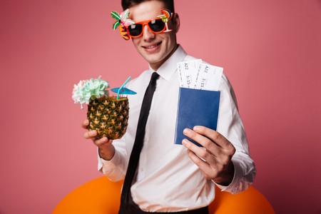 Cropped photo of excited happy young businessman with rubber ring standing isolated holding passport with tickets and cocktail. Looking at camera. Focus on passport.の写真素材