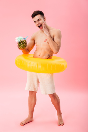Full length image of Happy naked man in shorts with inflatable ring holding cocktail and looking at the camera over pink backgroundの写真素材
