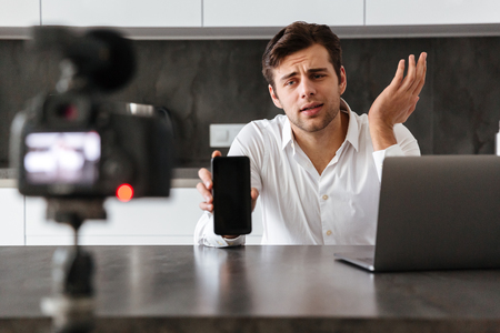 Handsome young man filming his video blog episode about new tech devices while sitting at the kitchen table and showing blank screen mobile phoneの写真素材