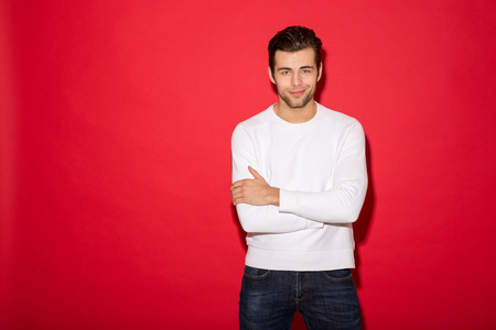 Cool smiling man in sweater looking at the camera with crossed arms over red backgroundの写真素材