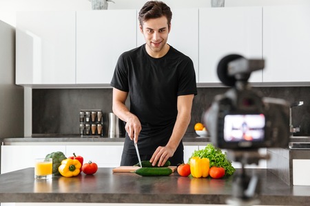 Handsome young man filming his video blog episode about healthy food cooking while standing at the kitchenの写真素材