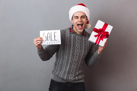 Image of young happy man wearing christmas santa hat standing isolated over grey wall holding paper with sale text and gift box. Looking camera.の写真素材