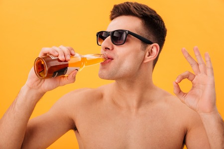 Close up view of Smiling naked man in sunglasses drinking beer and showing ok sign over yellow backgroundの写真素材