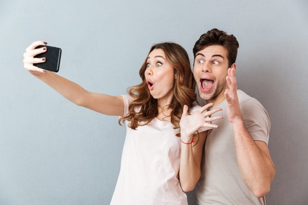 Portrait of an excited young couple standing and taking a selfie over gray wallの写真素材