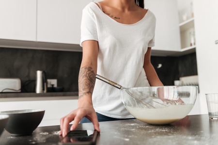 Cropped photo of amazing young woman standing at the kitchen in home cooking using mobile phone.の写真素材
