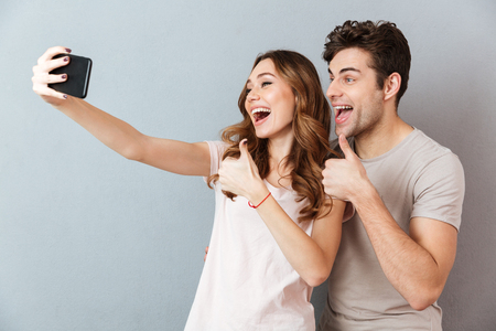 Portrait of a cheerful young couple showing thumbs up gesture while standing and taking a selfie over gray wallの写真素材