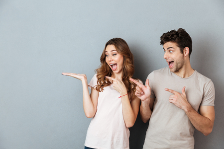 Portrait of a happy casual couple standing and pointing fingers away at copy space isolated over gray wall backgroundの写真素材