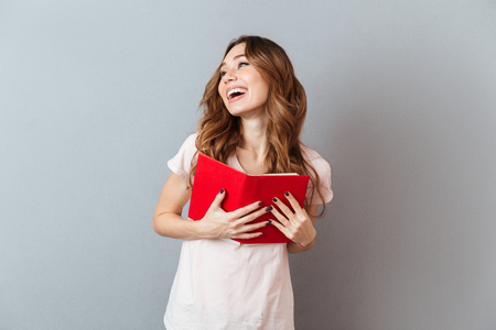 Portrait of a pretty laughing girl holding book and looking away isolated over gray wall backgroundの写真素材