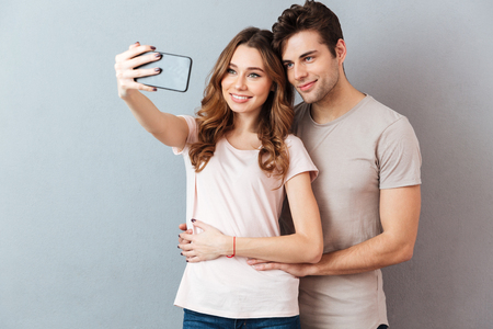 Portrait of a smiling young couple hugging while standing and taking a selfie over gray wallの写真素材