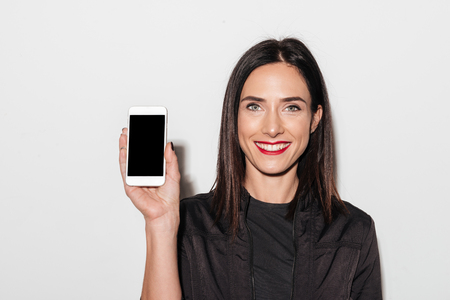 Image of young cheerful woman with red lips standing isolated over white wall. Looking camera showing display of mobile phone.の写真素材