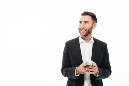 Portrait of a smiling young man holding mobile phone and looking away at copy space isolated over...