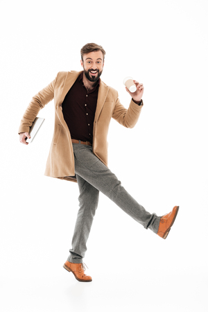 Full length portrait of a cheery young bearded guy dressed in coat holding laptop computer while walking and drinking coffee isolated over white backgroundの写真素材