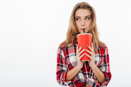 Portrait of a young cute girl in plaid shirt drinking from a plastic cup and looking away isolated over white backgroundの写真素材