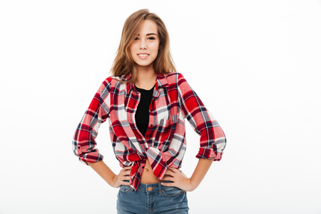 Portrait of a smiling casual girl in plaid shirt standing with arms on hips and looking at camera isolated over white backgroundの写真素材