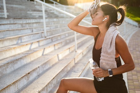 Portrait of a tired fitness girl with towel holding water bottle while resting after work out outdoorsの写真素材