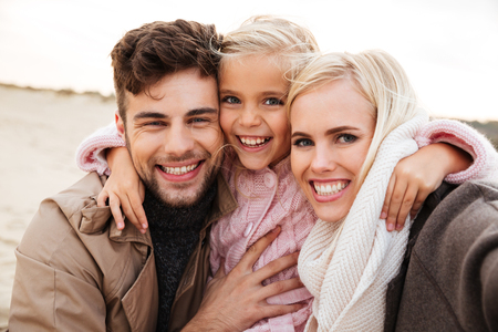 Portrait of a cheerful family with a little daughter taking a selfie together while standing at the beachの写真素材