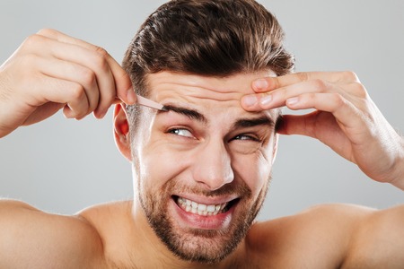 Beauty portrait of a handsome man plucking eyebrows with tweezers isolated over gray backgroundの写真素材