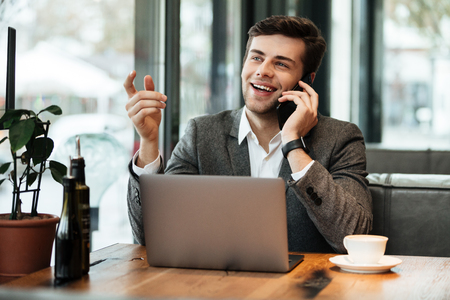 Happy business man sitting by the table in cafe with laptop computer while talking by smartphoneの写真素材