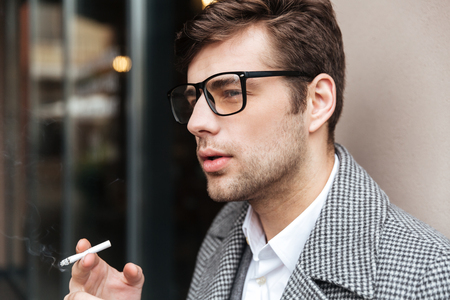Close up side view of Serious business man in eyeglasses and coat smoking cigarette outdoorsの写真素材