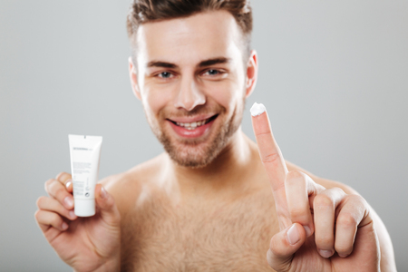 Beauty portrait of a happy half naked man applying face cream isolated over gray backgroundの写真素材