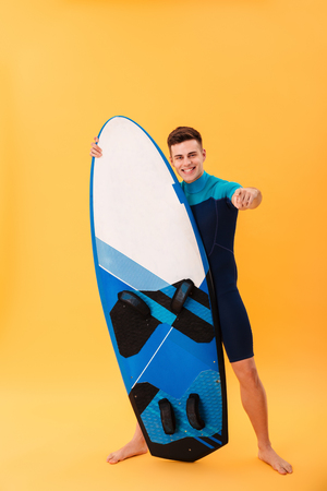 Full length portrait of confident smiling man in swimsuit pointing with finger on you while standing and holding surfboard, isolated over yellow backgroundの写真素材