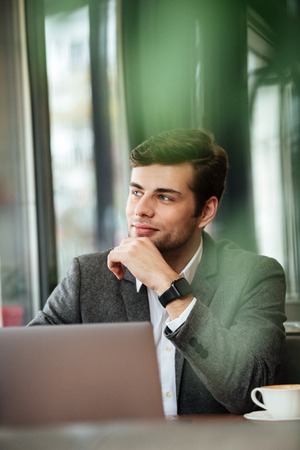 Vertical image of Pensive business man sitting by the table in cafe with laptop computer and looking...
